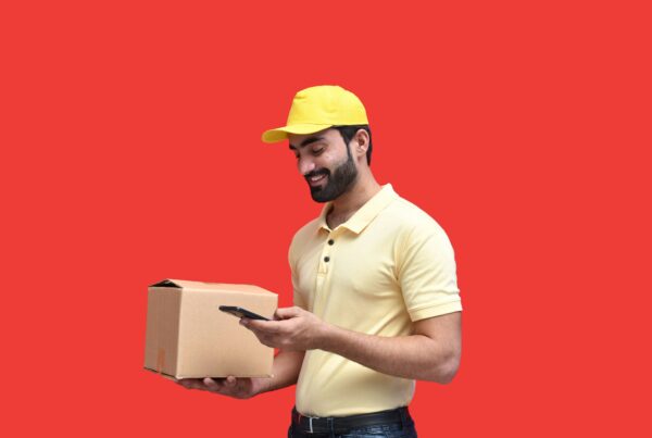 Lieferbote in gelber Uniform scannt ein Paket mit dem Smartphone vor rotem Hintergrund